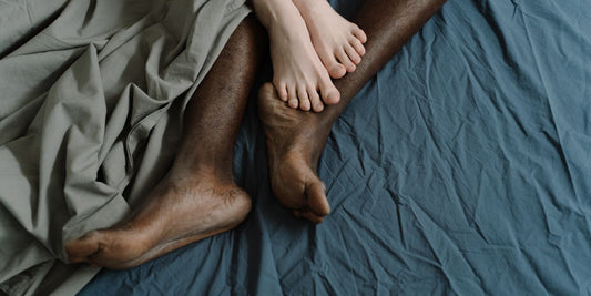 Pies de una pareja entrelazados sobre sábanas: guía para elegir un arnés sexual